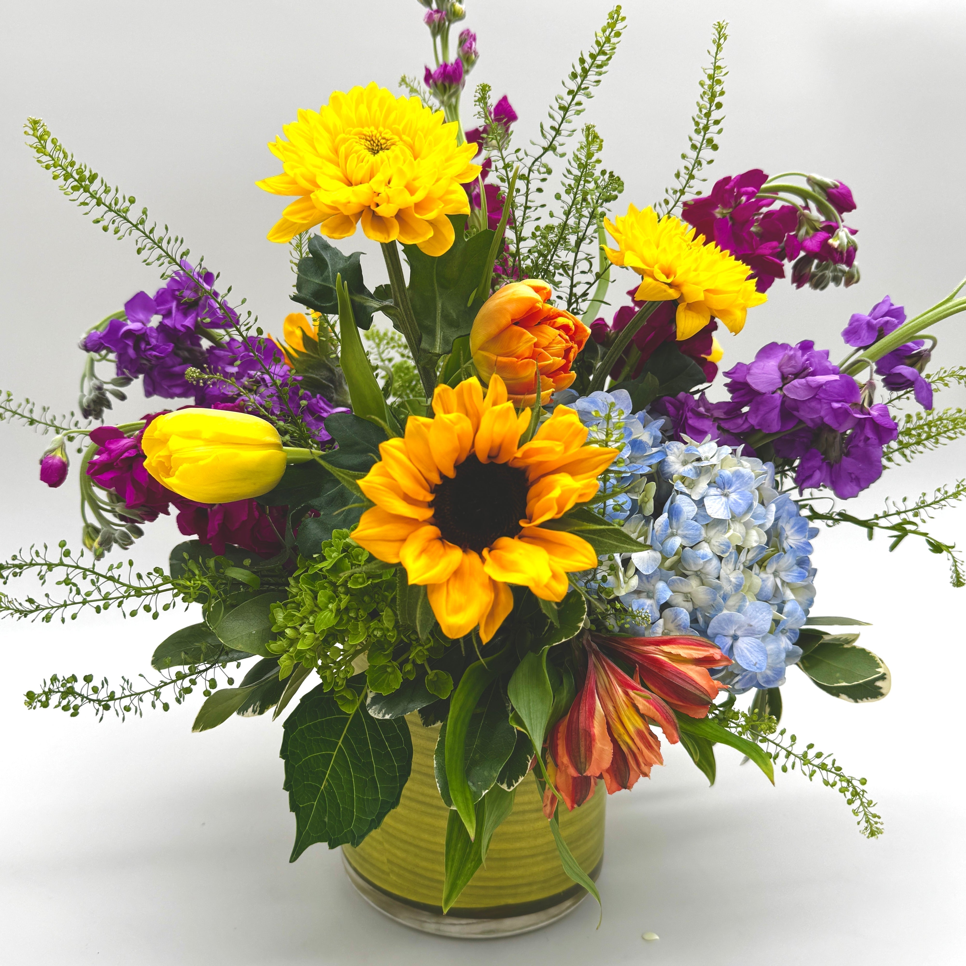 “Sunny Side Up” floral arrangement featuring sunflowers, yellow tulips, blue hydrangea, purple stock, alstroemeria, and seasonal blooms in a modern green glass vase, designed by floral designer Julie Davis at Blooms by Dragonfly.